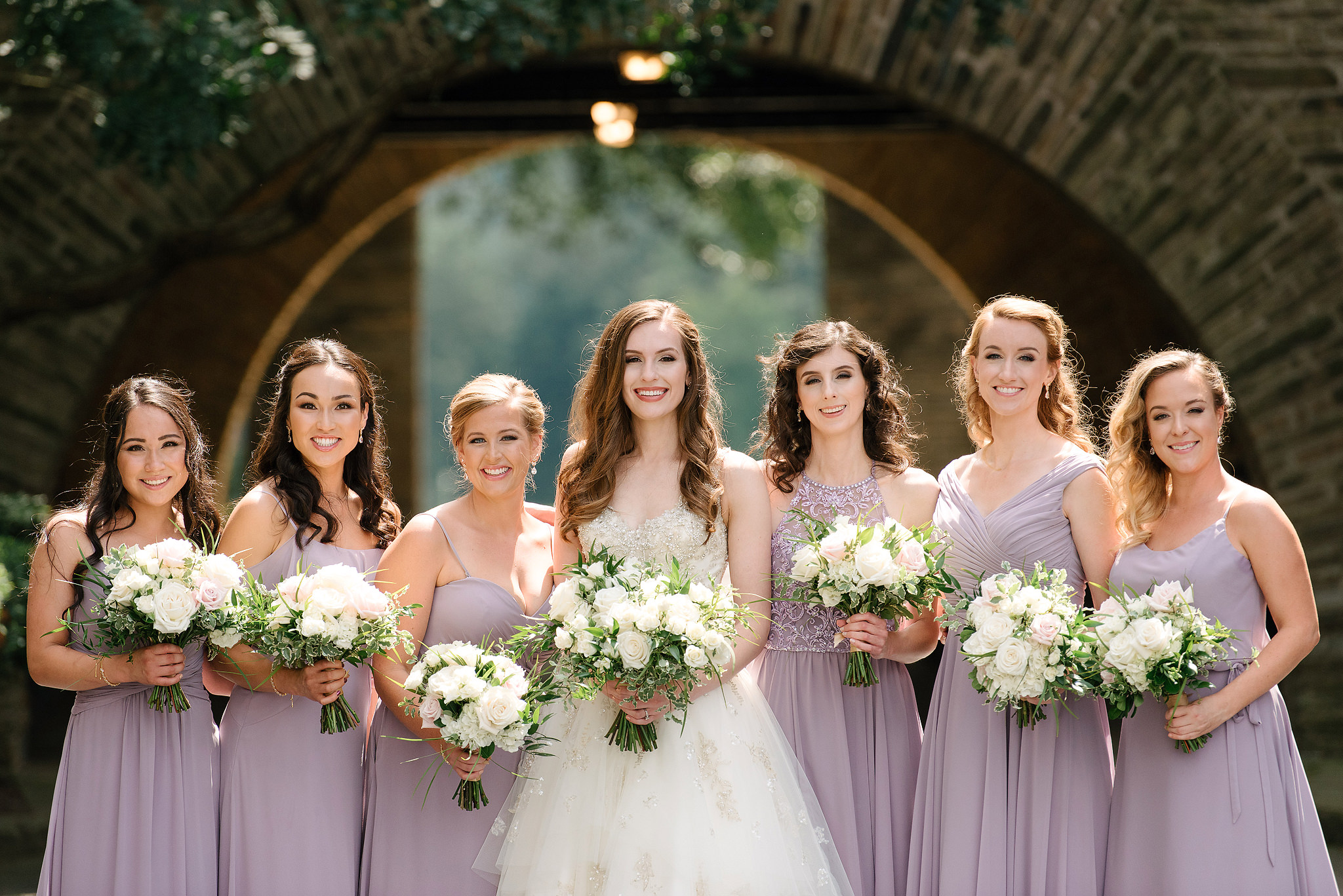 Bride with bridesmaids in lavender dresses beneath a stone arch